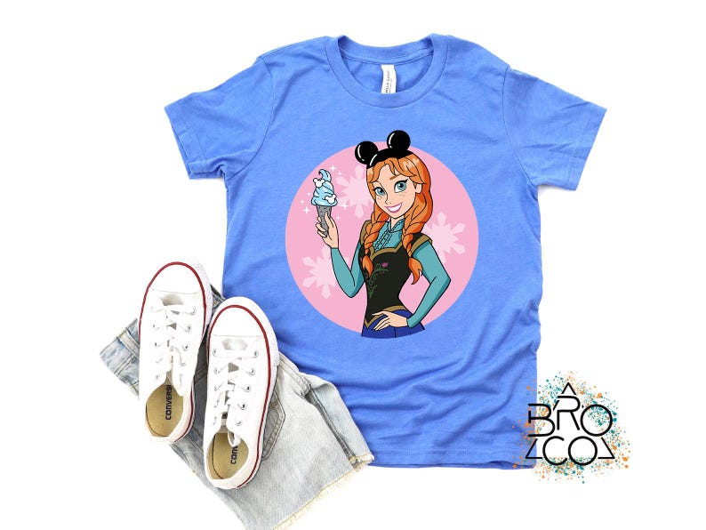 May include: A light blue t-shirt featuring a cartoon image of Anna from Frozen, holding an ice cream cone. The design is set within a pink circle with floral accents. White sneakers and denim shorts are placed below the shirt.