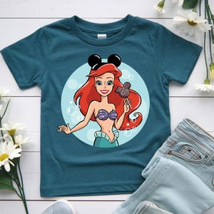 May include: Teal t-shirt featuring a cartoon illustration of a mermaid with red hair, a purple seashell bra, and a teal skirt. The mermaid is holding a Mickey Mouse-shaped lollipop. The shirt has a round neck and short sleeves.