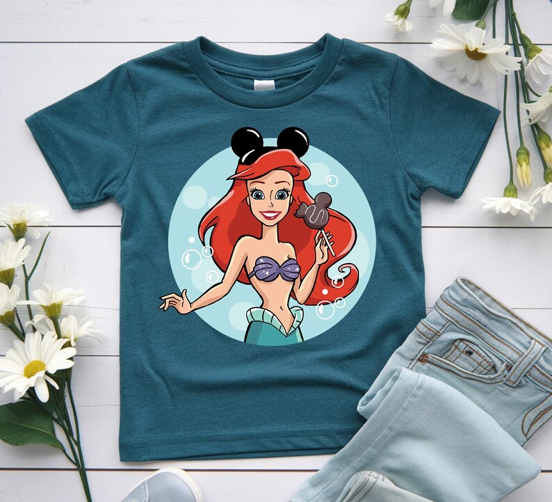 May include: Teal t-shirt featuring a cartoon illustration of a mermaid with red hair, a purple seashell bra, and a teal skirt. The mermaid is holding a Mickey Mouse-shaped lollipop. The shirt has a round neck and short sleeves.