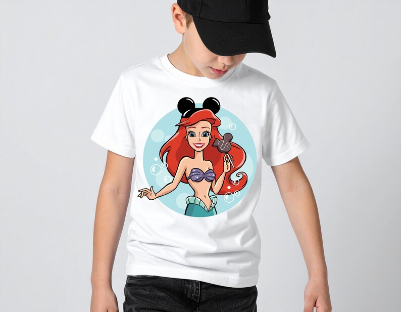 May include: White t-shirt featuring a cartoon illustration of Ariel from The Little Mermaid, with Mickey Mouse ears. Ariel has red hair, a purple seashell bra, and a teal tail. She is holding a Mickey Mouse-shaped lollipop. Bubbles surround her.