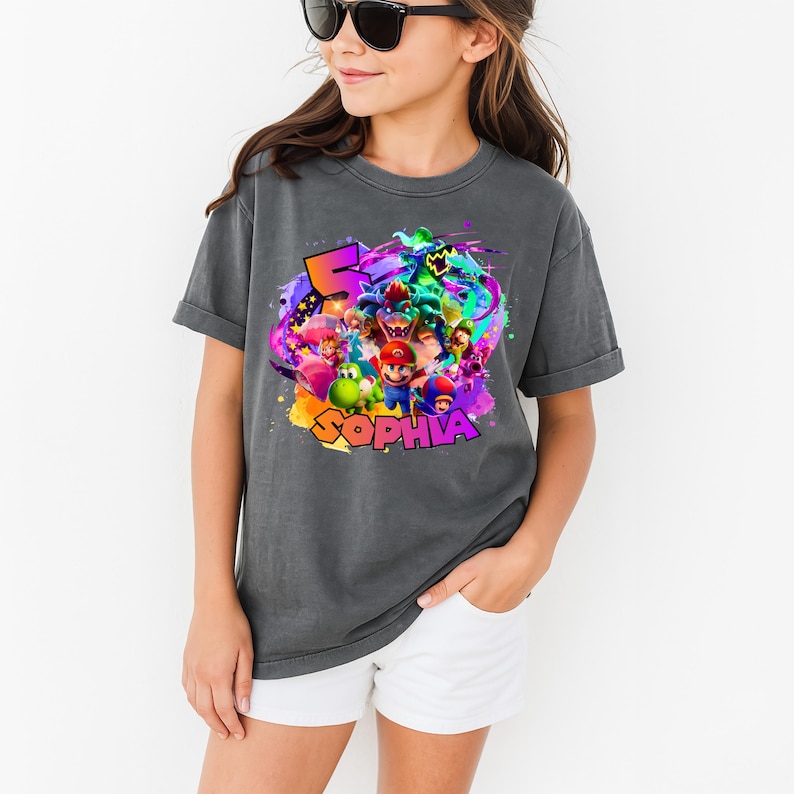 Puede incluir: Una camiseta gris carb&oacute;n de manga corta con un estampado gr&aacute;fico colorido de varios personajes de videojuegos, incluidos Mario, Luigi, Yoshi y Bowser. El dise&ntilde;o incluye un n&uacute;mero 5 grande y el nombre SOPHIA en letras gruesas y cuadradas.