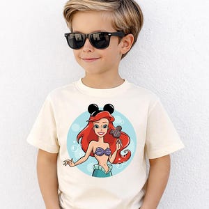 May include: A cream-colored t-shirt featuring a cartoon illustration of Ariel from The Little Mermaid. Ariel has red hair, a purple seashell top, and a blue skirt. She is holding a Mickey Mouse-shaped lollipop. The shirt also has black Mickey Mouse ears.