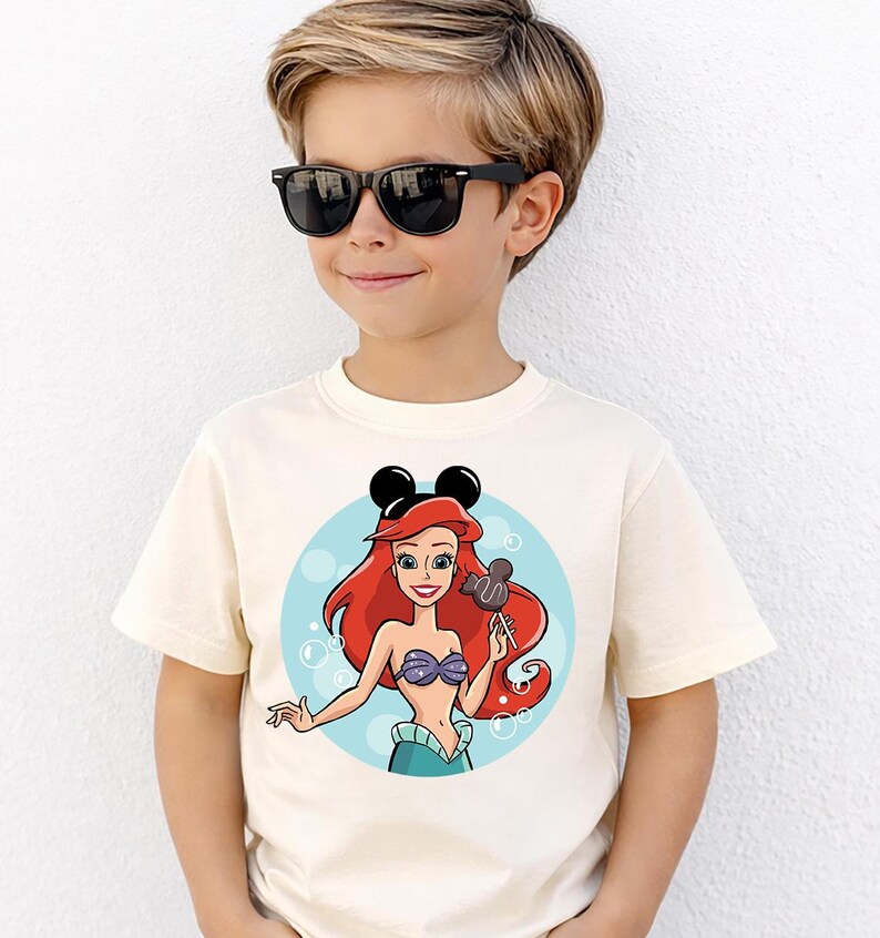 May include: A cream-colored t-shirt featuring a cartoon illustration of Ariel from The Little Mermaid. Ariel has red hair, a purple seashell top, and a blue skirt. She is holding a Mickey Mouse-shaped lollipop. The shirt also has black Mickey Mouse ears.