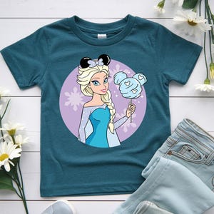 May include: Teal t-shirt featuring a cartoon illustration of a princess with blonde hair, wearing a blue dress and black mouse ears. She holds a blue cotton candy shaped like Mickey Mouse. A purple circle with snowflakes is behind her.
