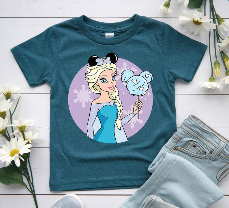 May include: Teal t-shirt featuring a cartoon illustration of a princess with blonde hair, wearing a blue dress and black mouse ears. She holds a blue cotton candy shaped like Mickey Mouse. A purple circle with snowflakes is behind her.