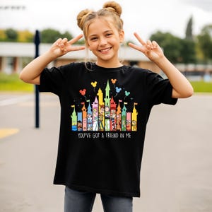 May include: Black t-shirt featuring a colorful castle design with cartoon characters and balloons. The text "YOU'VE GOT A FRIEND IN ME" is printed below the castle. The shirt is worn by a person.