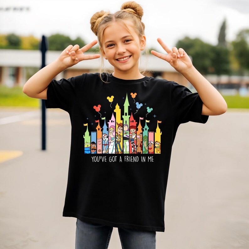 May include: Black t-shirt featuring a colorful castle design with cartoon characters and balloons. The text "YOU'VE GOT A FRIEND IN ME" is printed below the castle. The shirt is worn by a person.