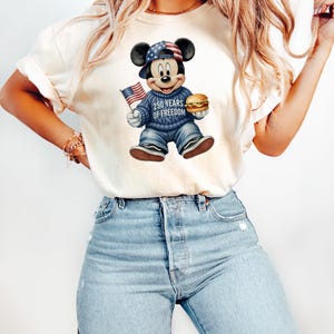 May include: Cream-colored t-shirt featuring a graphic of Mickey Mouse in a patriotic outfit. Mickey wears a hat and sweater with American flag designs, holding a burger and a small American flag. The text "250 YEARS OF FREEDOM" is on the sweater.