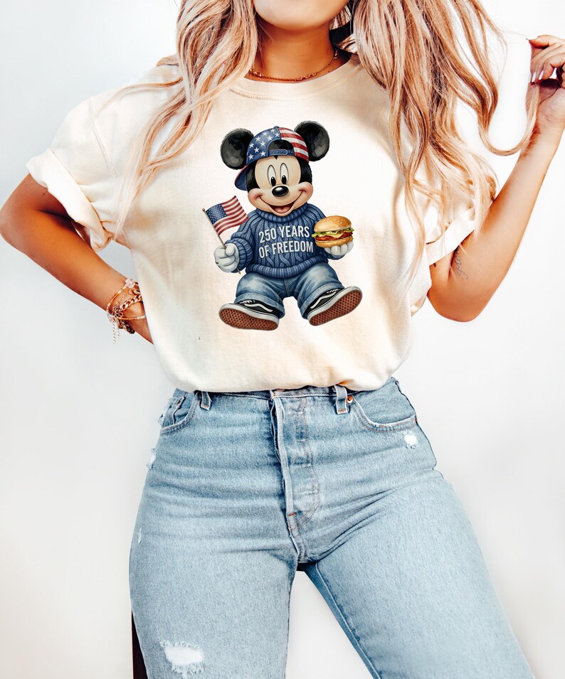 May include: Cream-colored t-shirt featuring a graphic of Mickey Mouse in a patriotic outfit. Mickey wears a hat and sweater with American flag designs, holding a burger and a small American flag. The text "250 YEARS OF FREEDOM" is on the sweater.
