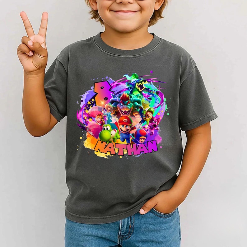Puede incluir: Una camiseta gris carb&oacute;n con un estampado gr&aacute;fico colorido de Mario, Luigi, Yoshi, Toad, Peach y Bowser. El dise&ntilde;o incluye un n&uacute;mero 8 grande y el nombre NATHAN en letras llamativas y vibrantes sobre un fondo de salpicaduras de pintura.