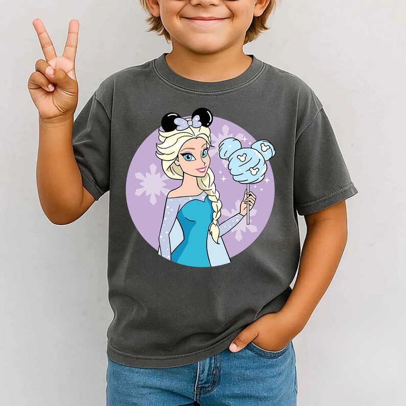 May include: Dark gray t-shirt featuring a cartoon image of Elsa from Frozen. Elsa is wearing a blue dress and holding a blue cotton candy. She has blonde hair and is wearing black mouse ears with a bow. The background is a purple circle with snowflakes.