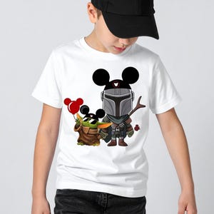 May include: White t-shirt featuring a cartoon illustration of The Mandalorian and Grogu, with Mickey Mouse ears. Grogu is holding red balloons. The Mandalorian is holding a spear. The shirt is worn by a person.