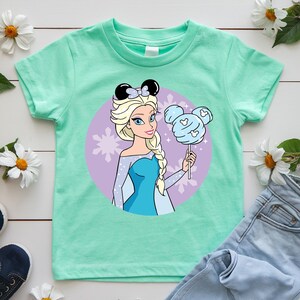 May include: A mint green t-shirt featuring a cartoon illustration of a princess with blonde hair and a blue dress. She is holding a blue cotton candy shaped like Mickey Mouse ears. The princess is set against a purple background with snowflakes.