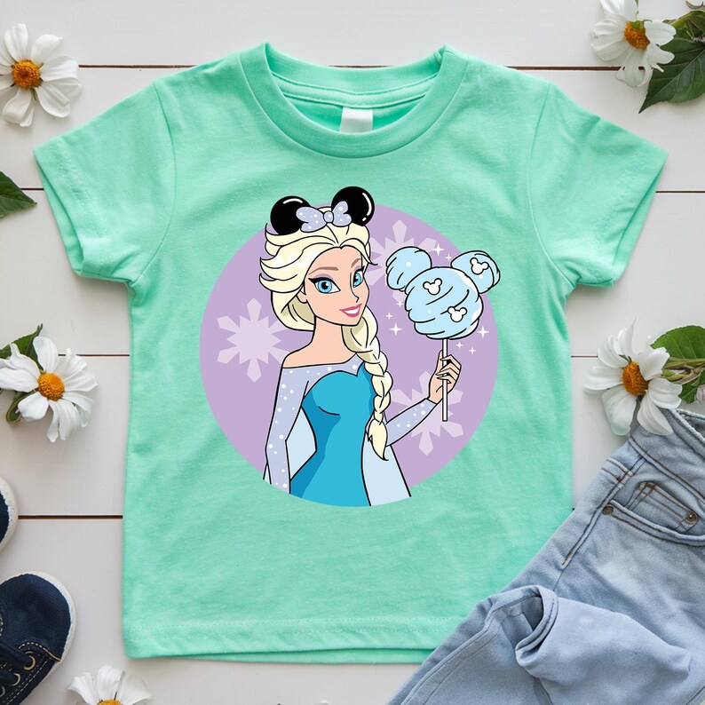 May include: A mint green t-shirt featuring a cartoon illustration of a princess with blonde hair and a blue dress. She is holding a blue cotton candy shaped like Mickey Mouse ears. The princess is set against a purple background with snowflakes.
