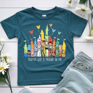 May include: Teal short-sleeved t-shirt featuring a colorful graphic of cartoon characters and castle towers. The text "YOU'VE GOT A FRIEND IN ME" is printed below the graphic. The shirt is paired with light blue jeans.