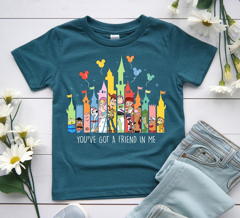 May include: Teal short-sleeved t-shirt featuring a colorful graphic of cartoon characters and castle towers. The text "YOU'VE GOT A FRIEND IN ME" is printed below the graphic. The shirt is paired with light blue jeans.