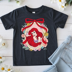 May include: Dark gray t-shirt featuring a cartoon illustration of a red-haired mermaid inside a floral frame with a red bow. The mermaid is wearing a purple top. The frame is surrounded by pearls and colorful flowers.