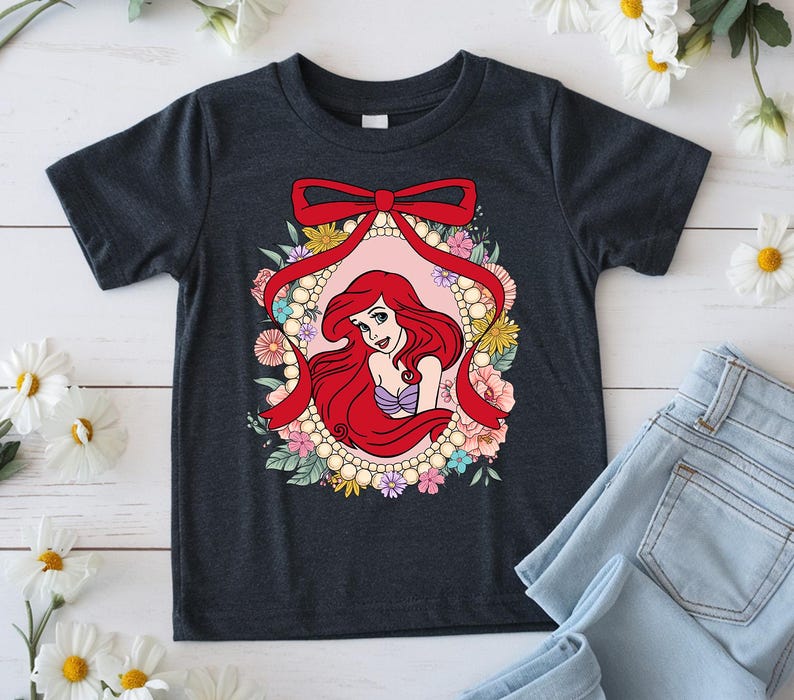 May include: Dark gray t-shirt featuring a cartoon illustration of a red-haired mermaid inside a floral frame with a red bow. The mermaid is wearing a purple top. The frame is surrounded by pearls and colorful flowers.