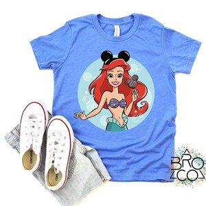 May include: A light blue t-shirt featuring a cartoon illustration of Ariel from The Little Mermaid, wearing Mickey Mouse ears and holding a Mickey Mouse-shaped lollipop. White sneakers and denim shorts are placed below the shirt.