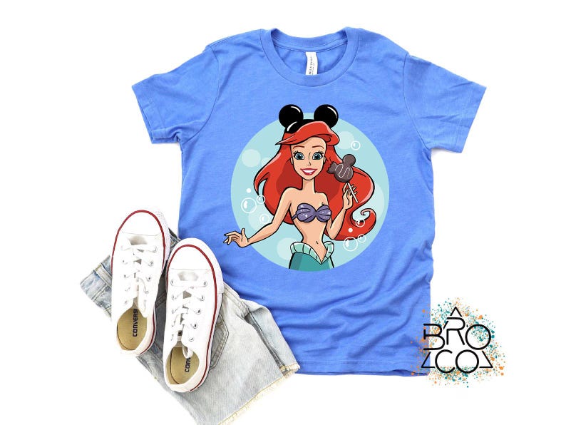 May include: A light blue t-shirt featuring a cartoon illustration of Ariel from The Little Mermaid, wearing Mickey Mouse ears and holding a Mickey Mouse-shaped lollipop. White sneakers and denim shorts are placed below the shirt.