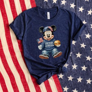 May include: Navy blue t-shirt featuring a cartoon Mickey Mouse design. Mickey is wearing a patriotic hat and sweater, holding a burger and an American flag. The text on the sweater reads "250 YEARS OF FREEDOM".