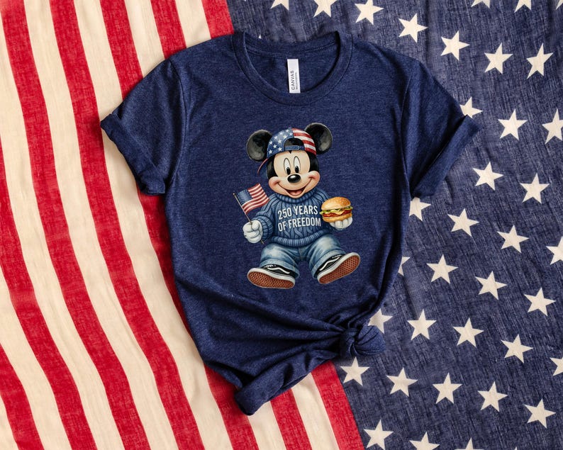 May include: Navy blue t-shirt featuring a cartoon Mickey Mouse design. Mickey is wearing a patriotic hat and sweater, holding a burger and an American flag. The text on the sweater reads "250 YEARS OF FREEDOM".