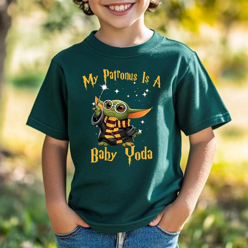 May include: Dark green t-shirt featuring the text "My Patronus Is A Baby Yoda." The graphic shows a cartoon Baby Yoda character with glasses, a wand, and a scarf. The design is in gold and white.