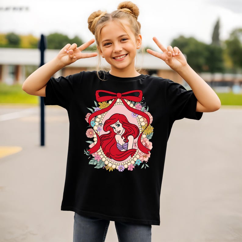 May include: Black t-shirt featuring a colorful graphic of a cartoon mermaid with red hair, a purple top, and a floral frame. The design includes a red bow and pearl accents. The shirt is worn by a person.