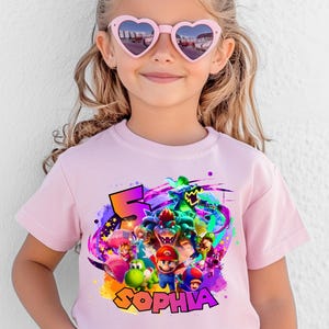 Puede incluir: Una camiseta de manga corta de color rosa claro con un estampado gr&aacute;fico colorido de Mario, Luigi, Princess Peach, Yoshi, Toad y Bowser. El dise&ntilde;o incluye un n&uacute;mero 5 grande y el nombre SOPHIA en letras amarillas negritas con borde negro.