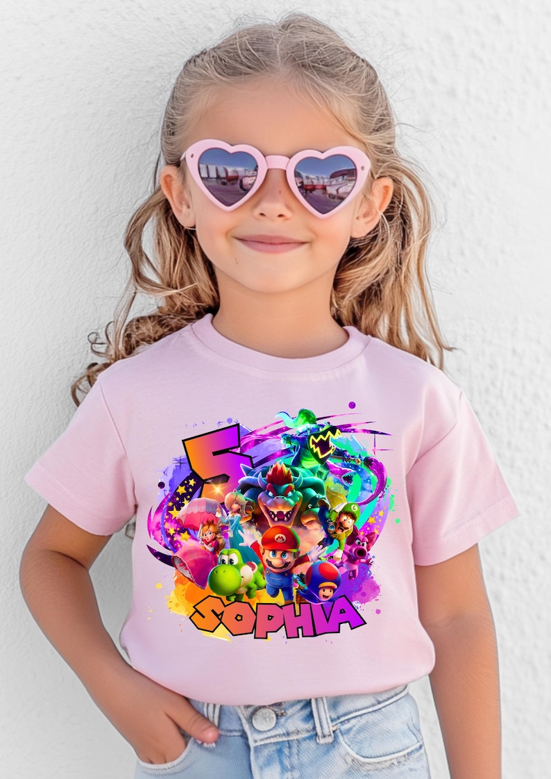 Puede incluir: Una camiseta de manga corta de color rosa claro con un estampado gr&aacute;fico colorido de Mario, Luigi, Princess Peach, Yoshi, Toad y Bowser. El dise&ntilde;o incluye un n&uacute;mero 5 grande y el nombre SOPHIA en letras amarillas negritas con borde negro.