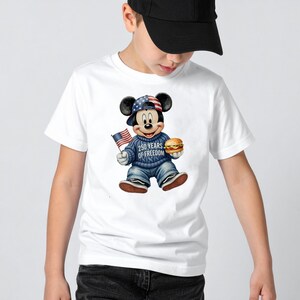 May include: White t-shirt featuring a cartoon Mickey Mouse design. Mickey is wearing a patriotic hat and sweater, holding an American flag and a hamburger. The sweater reads "250 Years of Freedom".