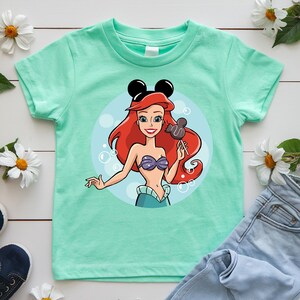 May include: Mint green t-shirt featuring a cartoon illustration of Ariel from The Little Mermaid. Ariel has red hair, a purple seashell bra, and is holding a chocolate-covered ice cream bar. Black Mickey Mouse ears are on her head.