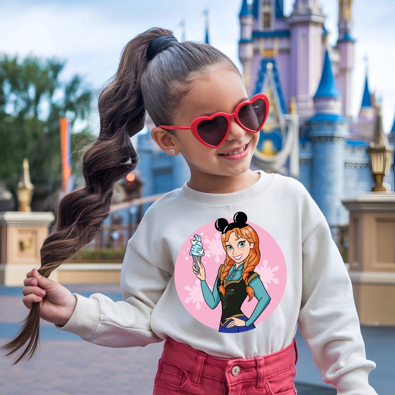 May include: A young person wearing a white sweatshirt featuring a cartoon image of Anna from Frozen holding an ice cream cone. The sweatshirt has a pink circle background with snowflakes. The person is also wearing red heart-shaped sunglasses and pink pants.
