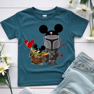 May include: Teal blue t-shirt featuring a cartoon illustration of The Mandalorian and Grogu, both wearing Mickey Mouse ears. Grogu holds a red balloon. The Mandalorian holds a twig and a red rose.