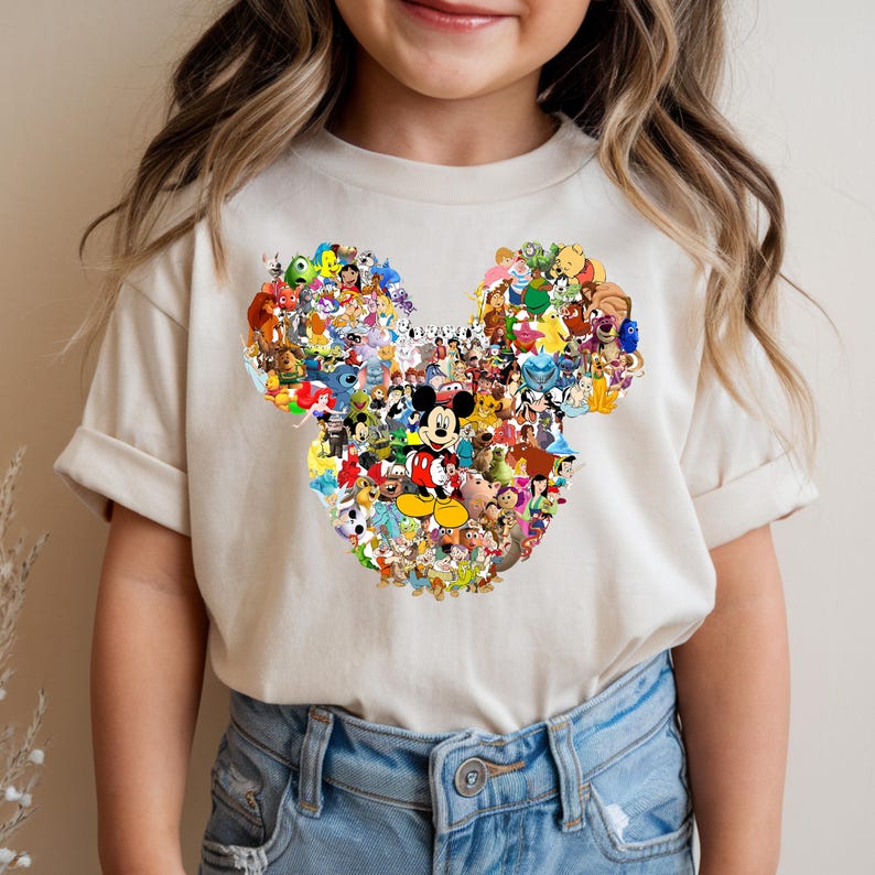 Puede incluir: Una camiseta color crema con un gran dise&ntilde;o de cabeza de Mickey Mouse lleno de varios personajes de Disney. El dise&ntilde;o incluye personajes de pel&iacute;culas cl&aacute;sicas y modernas de Disney, creando un gr&aacute;fico colorido y detallado.