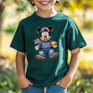 May include: Dark green t-shirt featuring a graphic of Mickey Mouse wearing a patriotic hat and sweater, holding an American flag and a burger. The text "250 YEARS OF FREEDOM" is on the sweater. The shirt is worn by a child.
