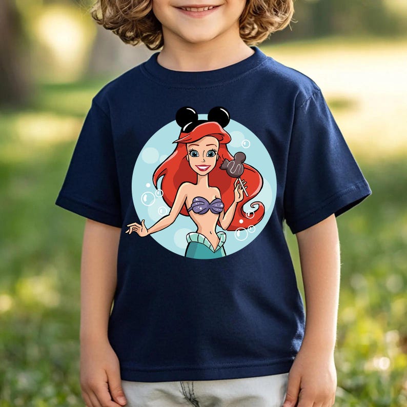 May include: Navy blue t-shirt featuring a cartoon illustration of Ariel from The Little Mermaid. Ariel has red hair, a purple seashell top, and a green tail. She is holding a Mickey Mouse-shaped lollipop and wearing Mickey Mouse ears.