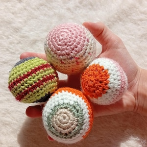 Op de afbeelding: Een hand houdt vier kleurrijke, gehaakte ballen vast. De ballen hebben verschillende kleuren, waaronder roze, oranje, groen en wit, met gestreepte en cirkelvormige patronen. De ballen lijken zacht en handgemaakt.