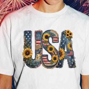 Puede incluir: Camiseta blanca con la palabra "USA" en un diseño patriótico. Las letras están decoradas con mezclilla, girasoles y la bandera estadounidense. La camiseta es de estilo casual.