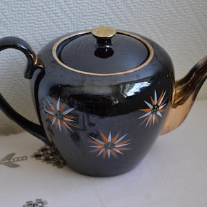 Może przedstawiać: Czarny ceramiczny czajnik ze złotym obramowaniem, uchwytem i wylewką. Czajnik ma pokrywkę ze złotym guzikiem i jest ozdobiony białymi, pomarańczowymi i złotymi wzorami w kształcie gwiazd. Czajnik stoi na białej powierzchni.