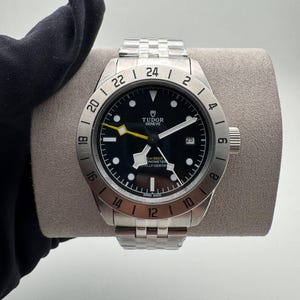 Può includere: Un orologio da polso in acciaio inossidabile con quadrante nero, indici e lancette bianche e lancetta dei secondi gialla. L'orologio ha un datario e una lunetta girevole. Anche il cinturino dell'orologio è in acciaio inossidabile.