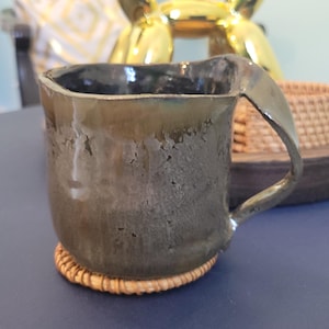 Könnte beinhalten: Eine handgefertigte Keramiktasse mit einer einzigartigen, strukturierten Oberfläche und einer dunklen, erdigen Farbe. Die Tasse hat einen markanten Griff und steht auf einem kleinen, runden gewebten Untersetzer. Der Innenraum ist dunkler.