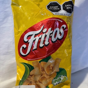 May include: A yellow bag of Fritos lime and salt flavored corn chips. The bag has a red oval with the word "Fritos" in white. The bag also has the words "Exceso Calorias" and "Exceso Sodio". The net weight is 6 oz (170 g).