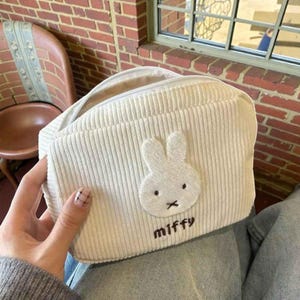 Peut inclure: Trousse de maquillage en velours côtelé crème avec une fermeture éclair et une poignée blanches. Elle présente un appliqué de lapin blanc avec des yeux noirs et une bouche en forme de "x" noire. Le mot "miffy" est brodé en noir.
