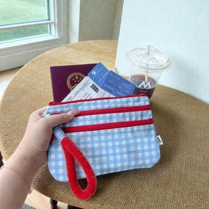 Puede incluir: Un estuche a cuadros azules y blancos con cremalleras rojas y una correa de muñeca roja. Un pasaporte y dos tarjetas de embarque son visibles en el fondo. El estuche se sostiene en una mano.