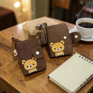 Pode incluir: Duas carteiras marrons com um desenho de urso de desenho animado, uma com bolinhas brancas. Um pequeno caderno bege e uma xícara de café estão em uma mesa de madeira. Uma bolsa bege com um chaveiro e uma bolinha fofa também está presente.