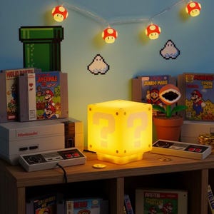 Puede incluir: Una lámpara de bloque amarillo con un signo de interrogación iluminado se encuentra sobre una superficie de madera, rodeada de cartuchos de juegos de Nintendo y una consola. Luces de cadena con forma de hongo y personajes pixelados decoran la pared azul.