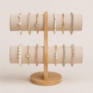 May include: A two-tiered wooden jewellery display stand holding twelve bracelets. The top tier features pearl, beaded, and gold-tone metal bangles. The bottom tier displays larger pearl strands, a gold chain with a round stone, and beaded bracelets.