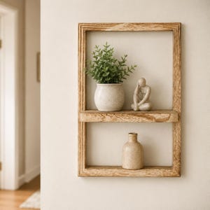 Pode incluir: Uma prateleira de parede retangular de madeira com dois níveis horizontais. A prateleira superior contém uma pequena planta verde em vaso e uma estatueta bege sentada. A prateleira inferior contém um vaso de cerâmica bege. Madeira com veios.