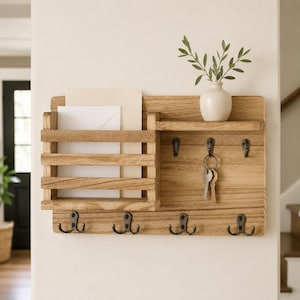 May include: A wall-mounted wooden organizer featuring a mail slot on the left, a small shelf on the right, and seven dark metal hooks. The mail slot holds two white envelopes, and the shelf displays a small white vase with green leaves.